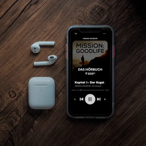 Smartphone Hörbuch-Player Mission: Goodlife neben AirPods und Ladecase auf Holz