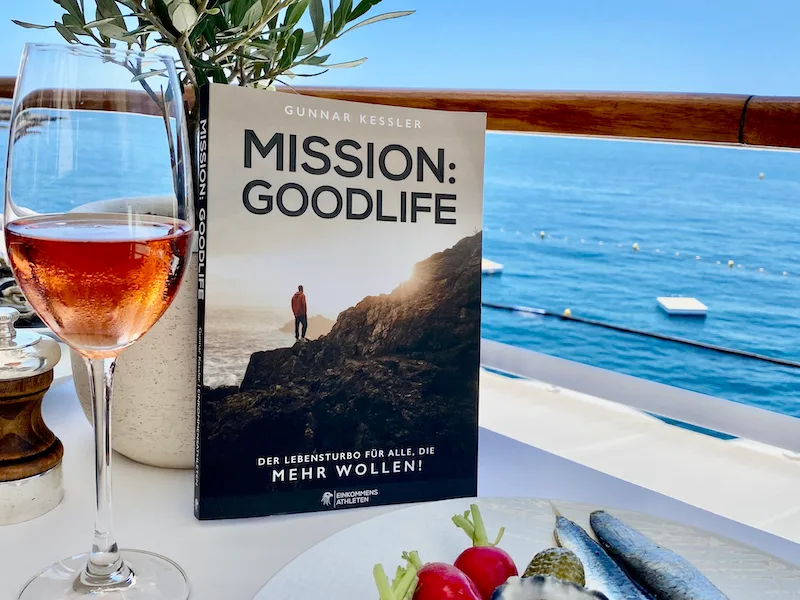 Mission: Goodlife Buch auf einem Tisch am Meer mit Rosé-Wein und Speisen