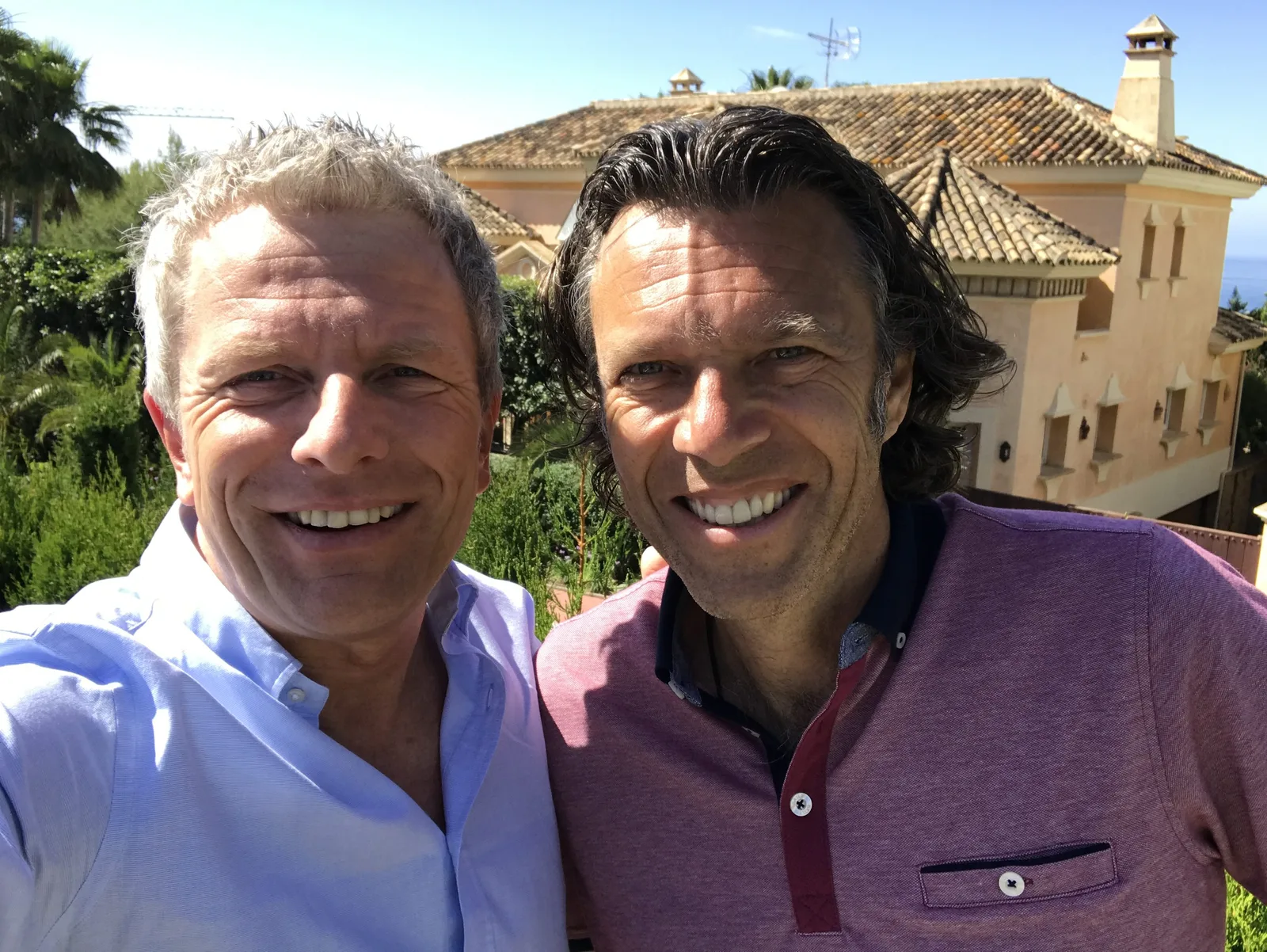 Urs Meier und Andreas Enrico Brell lächeln vor spanischem Herrenhof mit Palmen und Mittelmeer