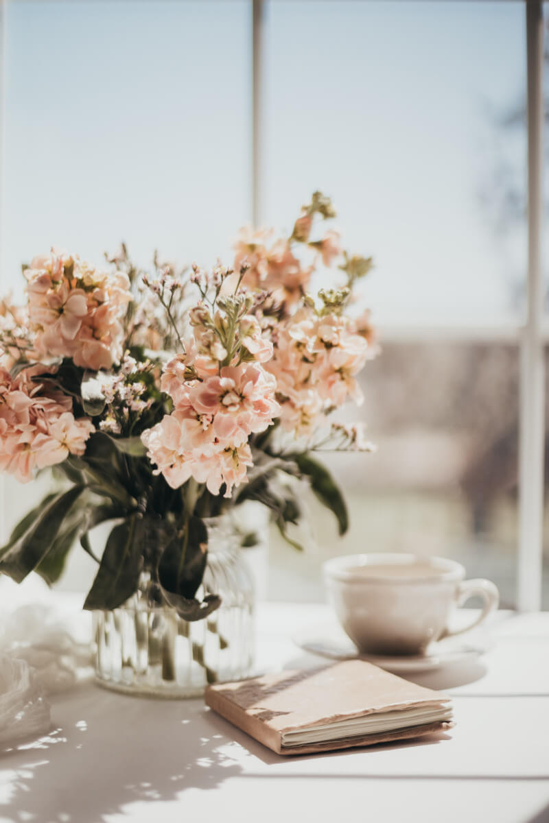 Stillleben mit Rosa Blumenstrauß in Glasvase, Tasse Kaffee und offenem Notizbuch am Fenster