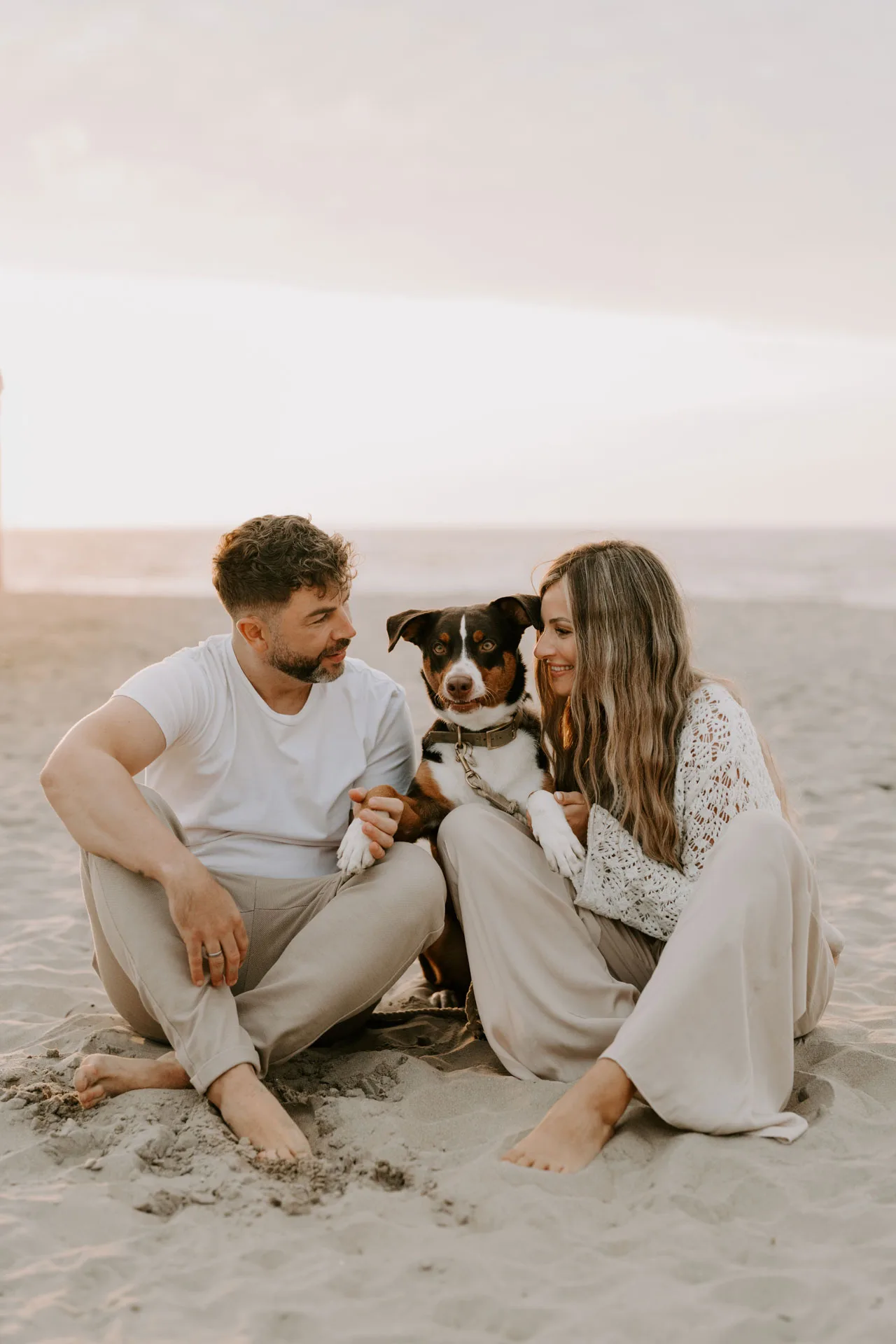 Paar sitzt mit braun-weißem Hund am Strand, entspannte Paarfotografie