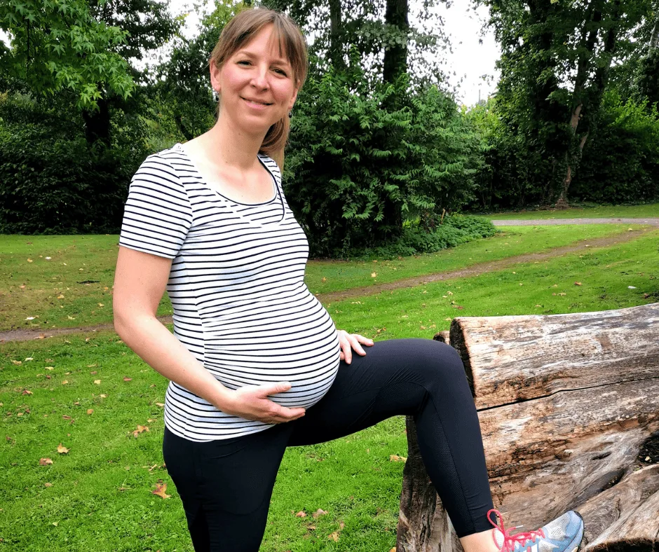 Schwangere Frau in gestreiftem Shirt posiert lächelnd im Park auf Baumstamm