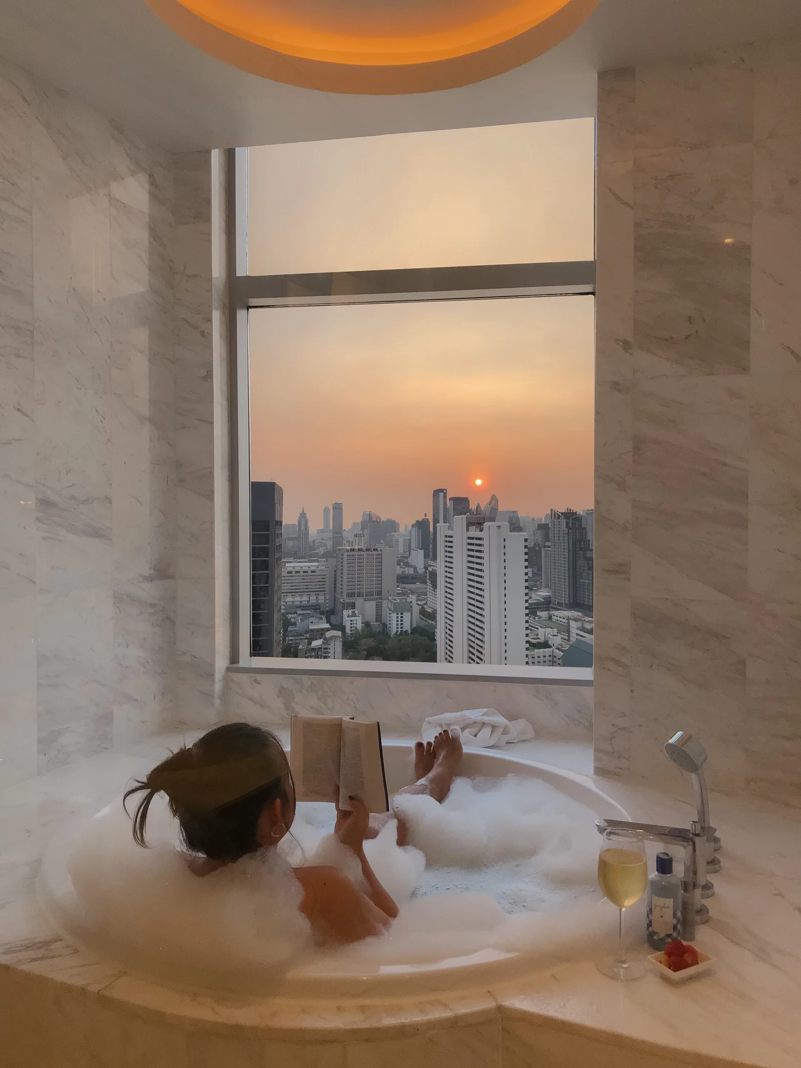 Frau liest in Luxus-Badewanne mit Blick auf Sonnenuntergang über Stadtskyline