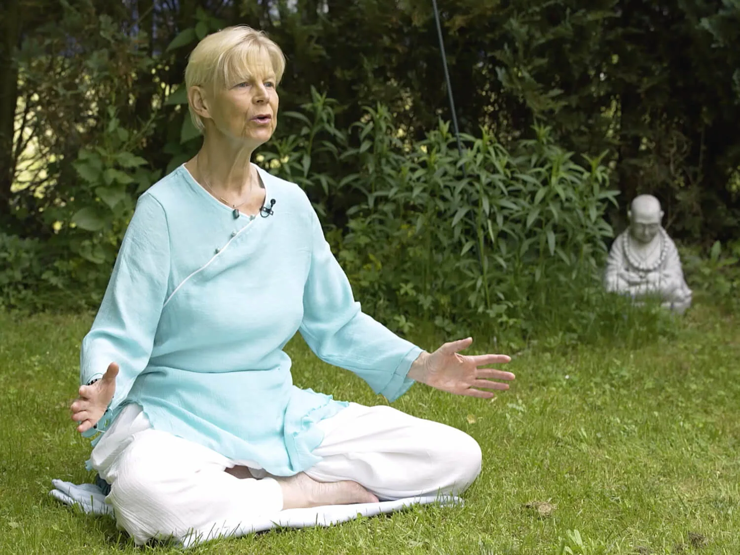 Nèizàng Ànmó – Qi-Gong-Videokurs von Annette Deinzer 5 Annette Deinzer sitzt im Qi-Gong-Lotussitz im Garten, türkisfarbenes Oberteil, weiße Hose