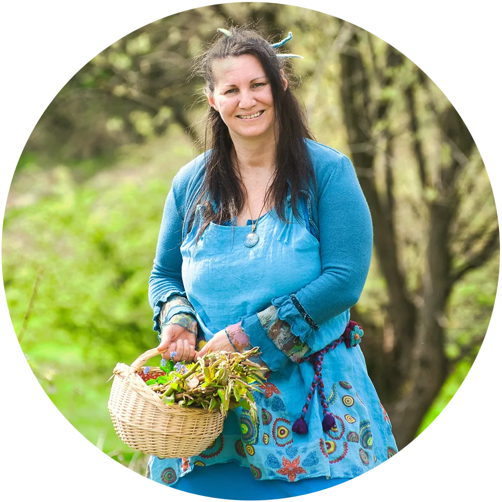 Sandra Valentin mit blauem Kleid und Körbchen voller Heilkräuter, steht lächelnd auf grüner Wiese mit Wald im Hintergrund