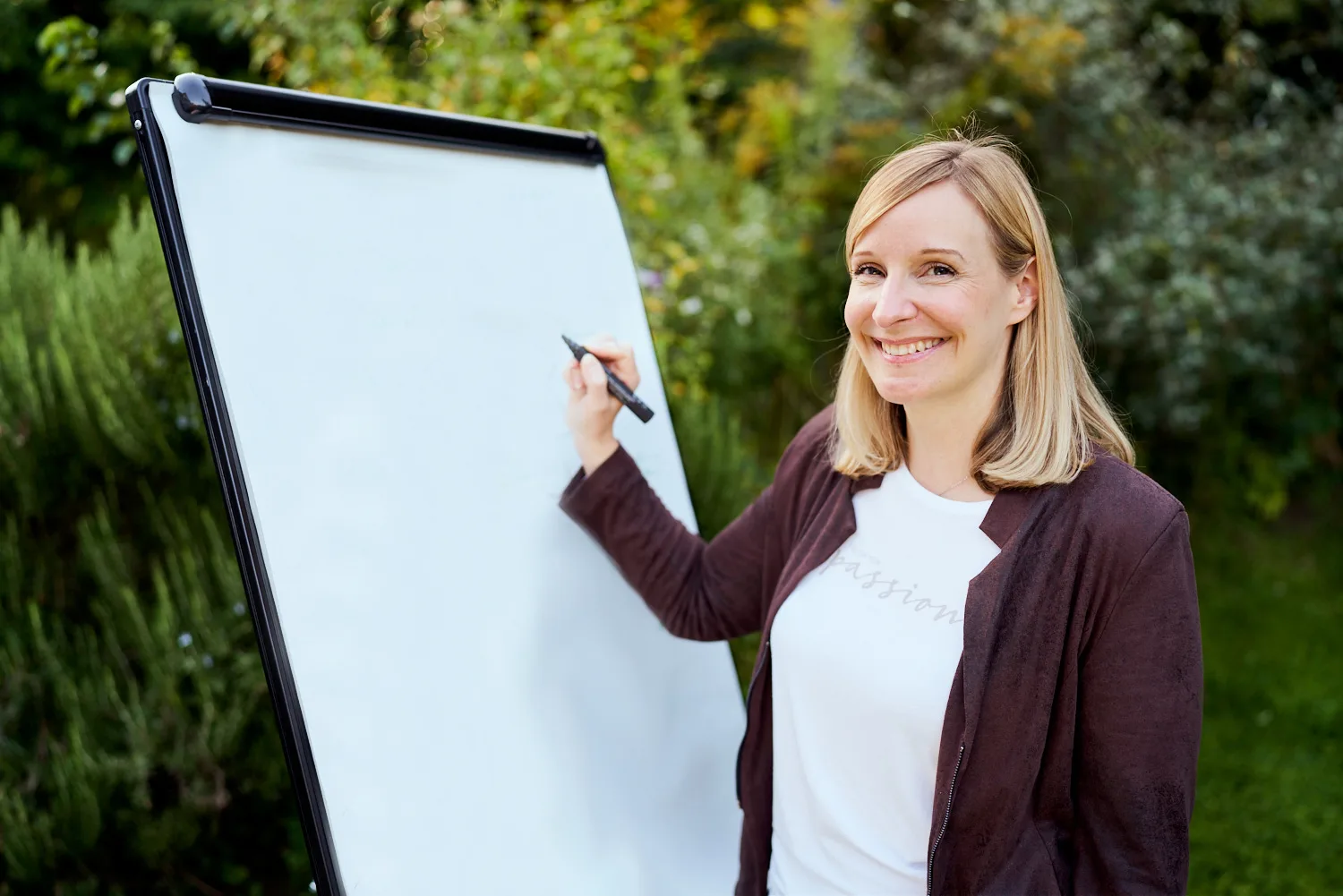 Frau mit blonden Haaren schreibt auf Flipchart vor grünem Buschwerk, trägt Strickjacke