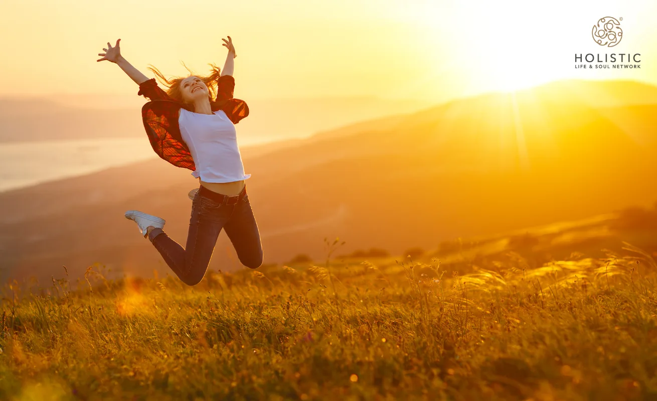Frau mit ausgebreiteten Armen springend auf feldiger Landschaft bei Sonnenuntergang, Holistic Logo