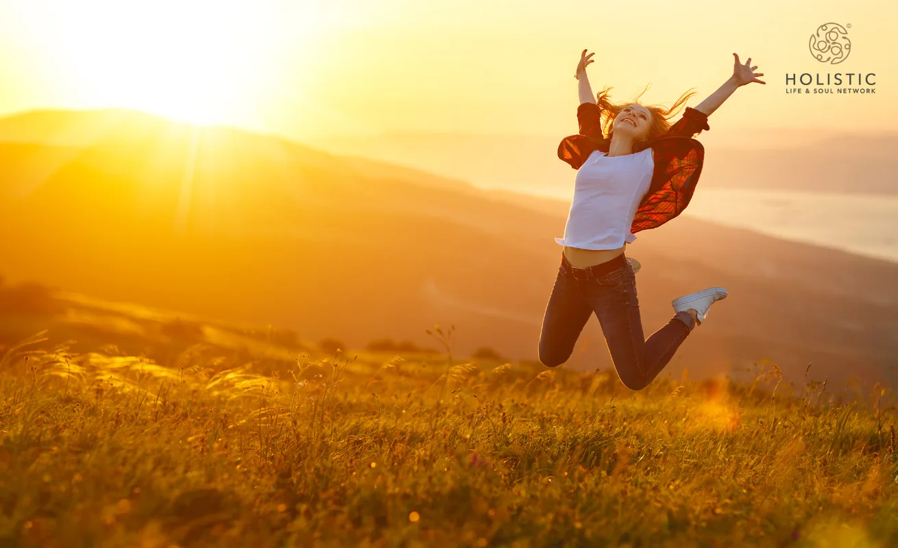 Frau springt freudig auf Wiese bei Sonnenaufgang mit ausgebreiteten Armen, Holistic Life & Soul Network Logo oben rechts