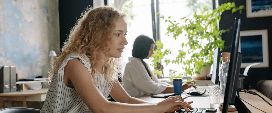 Zwei Frauen sitzen fokussiert am Schreibtisch vor ihren Computern im hellen Büro