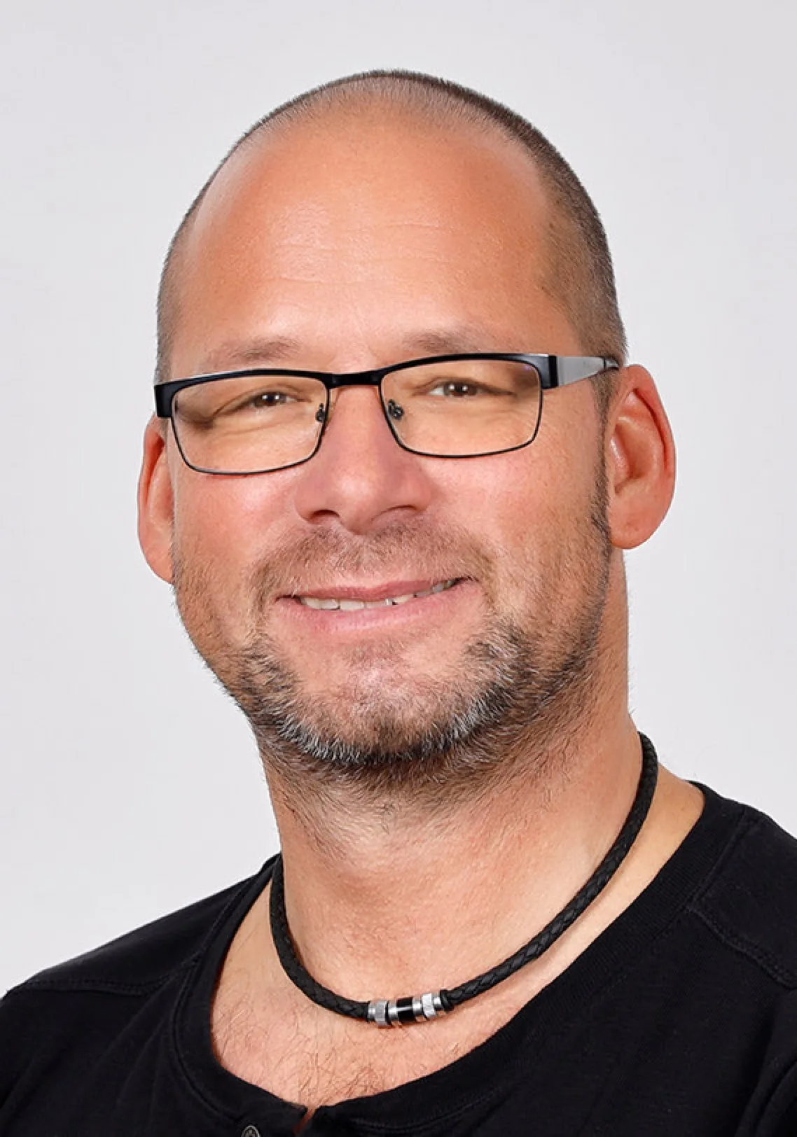 Thomas Seidl-Baresch, Mann mit Brille und Halskette in schwarzem T-Shirt vor hellem Hintergrund
