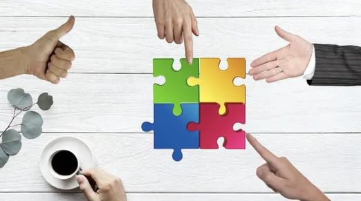 Onboarding – Kurs zur Mitarbeitereinarbeitung von Kristoffer Ditz 2 Vier Hände platzieren bunte Puzzle-Teile (grün, gelb, blau, rot) auf weißem Holztisch