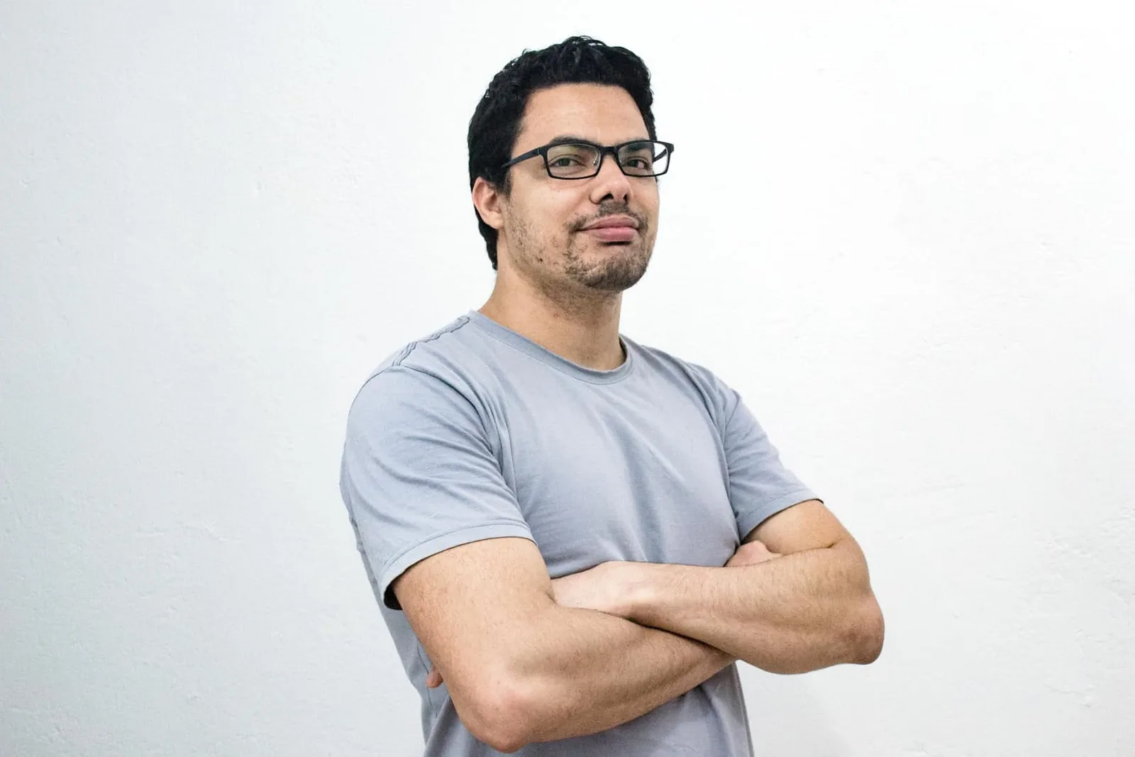 Mann mit verschränkten Armen, Brille und grauem T-Shirt blickt zur Seite vor weißem Hintergrund