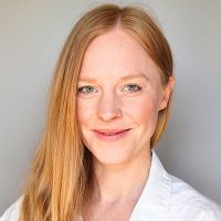 Svenja Adamek, Frau mit rötlichem Haar, weißes Oberteil, neutraler Hintergrund