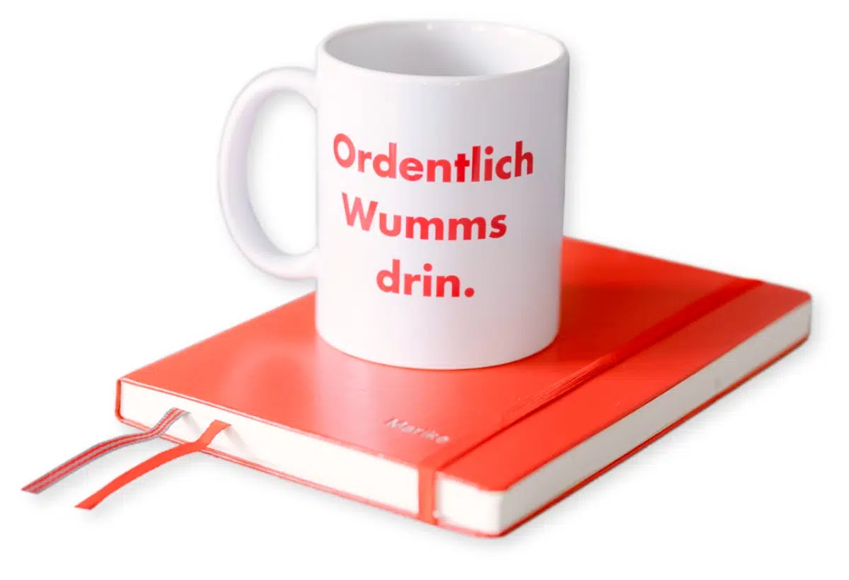 Weiße Tasse mit Text Ordentlich Wumms drin auf rotem Notizbuch