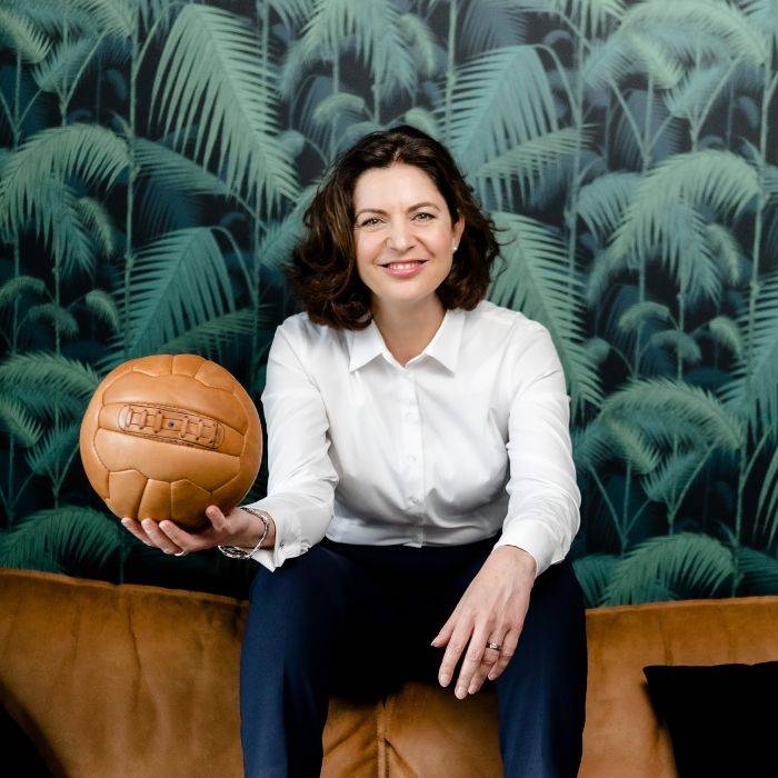 Bianca Traber mit brauner Bluse sitzt mit braunem Lederball vor tropischem Hintergrund