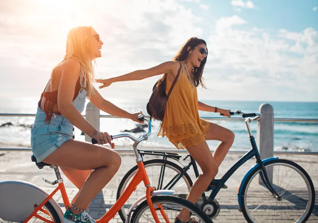 Zwei Frauen fahren lachend auf Fahrrädern an sonnigem Strand entlang