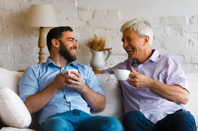 Zwei Männer unterschiedlichen Alters sitzen auf Sofa, trinken Kaffee und sprechen