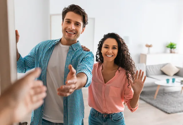 Zwei Personen öffnen Wohnungstür und begrüßen Besucher herzlich winkend