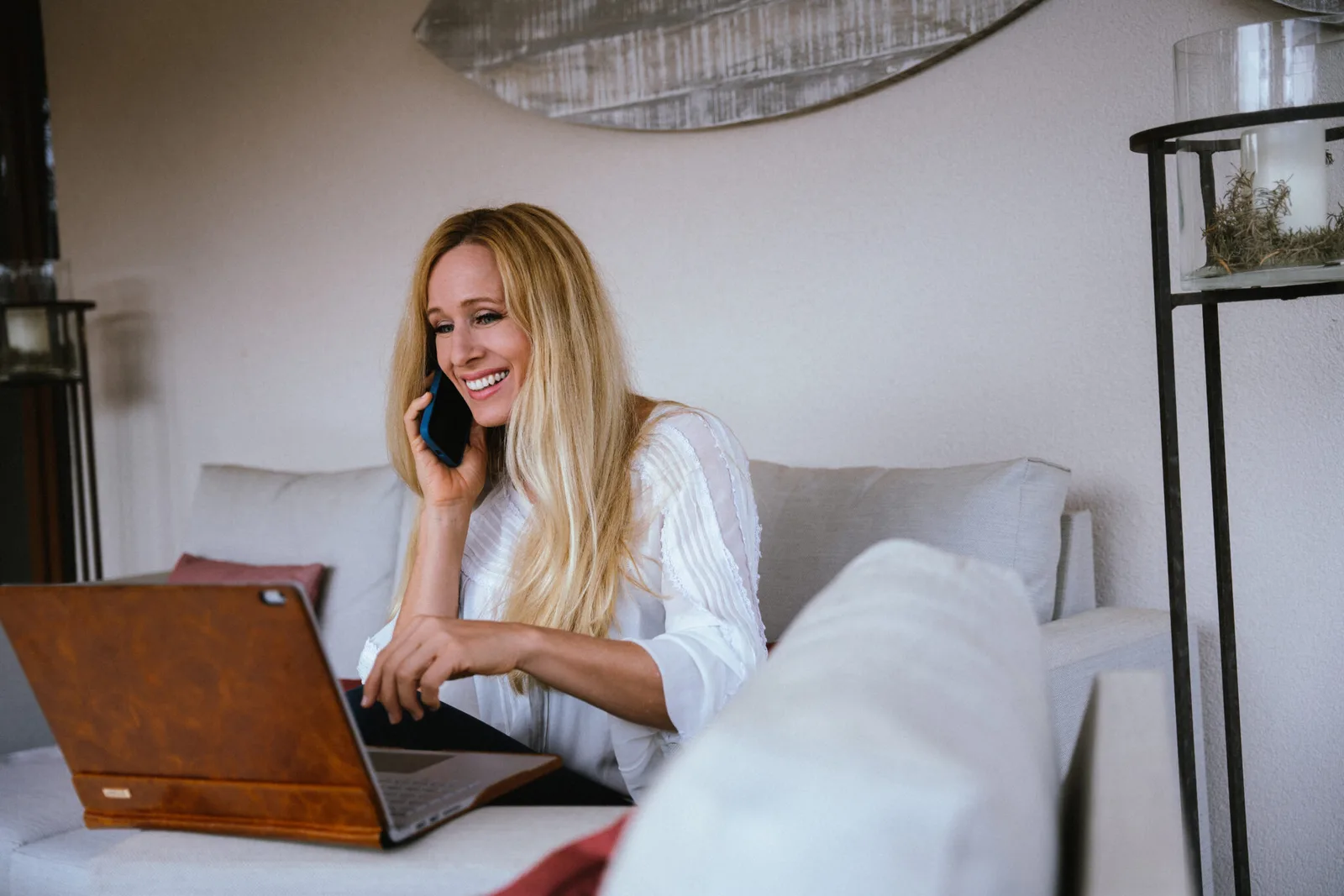 Frau mit blondem Haar sitzt auf Bett mit Laptop und telefoniert, modernes Schlafzimmer