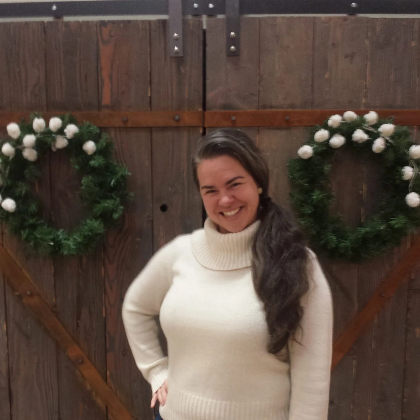 Frau in cremefarbenem Rollkragenpullover vor Holztor mit weißen Weihnachtskränzen