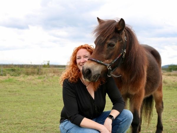 Frau mit roten Haaren kniet neben braunem Pferd auf Wiese
