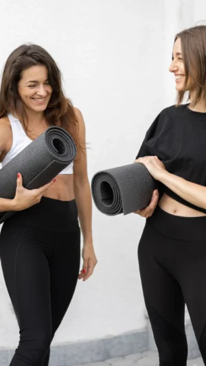 Zwei Frauen in Sportkleidung halten Yoga-Matten, lächeln sich an
