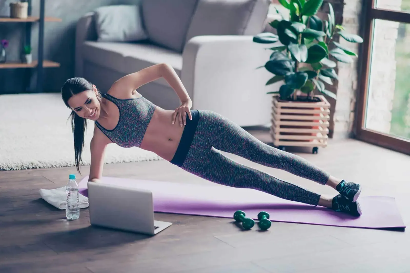 Junge Frau in Sportkleidung macht Seitstütz auf Trainingsmatte mit Laptop