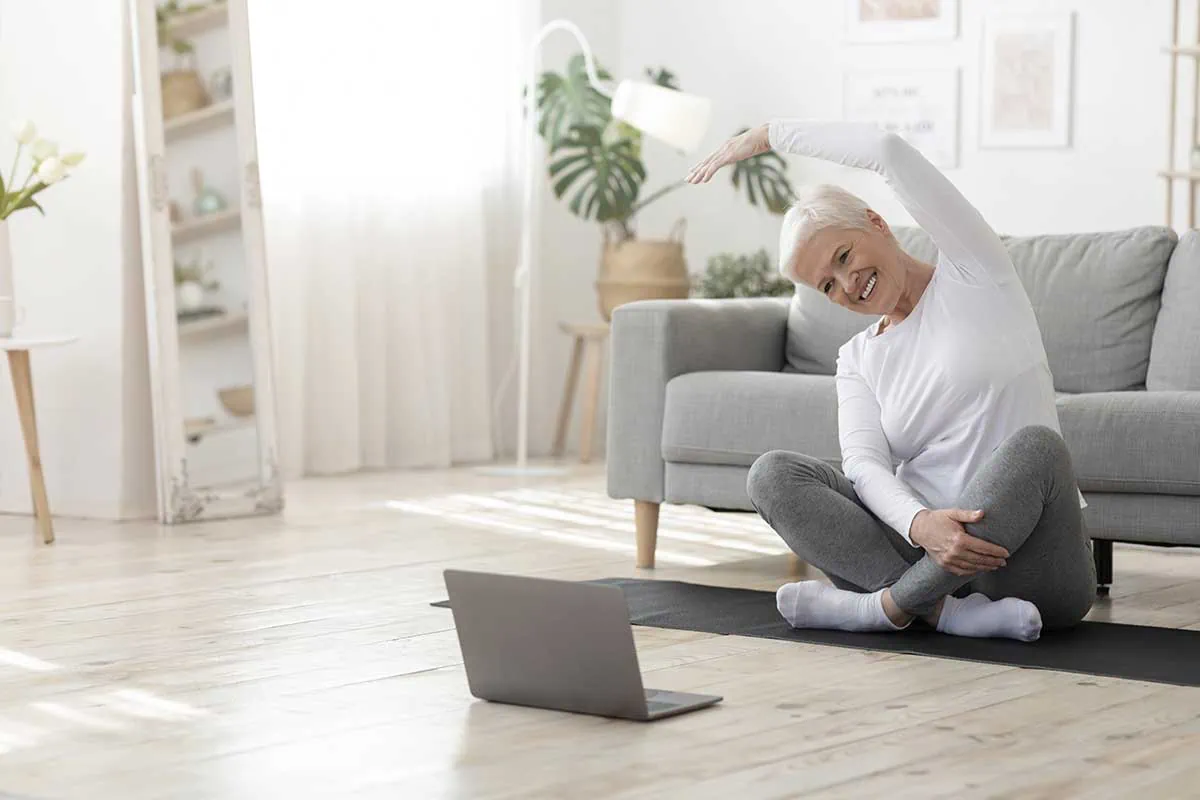 Ältere Frau macht Pilates-Stretching auf Yogamatte vor Laptop in hellem Wohnzimmer