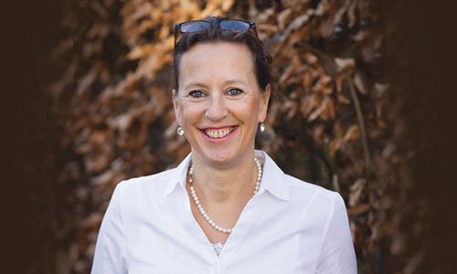 Porträt Antje Heimsoeth, Frau mit brünetten Haaren, weißer Bluse, Perlenkettern und Sonnenbrille im Haar vor braunem Naturhin