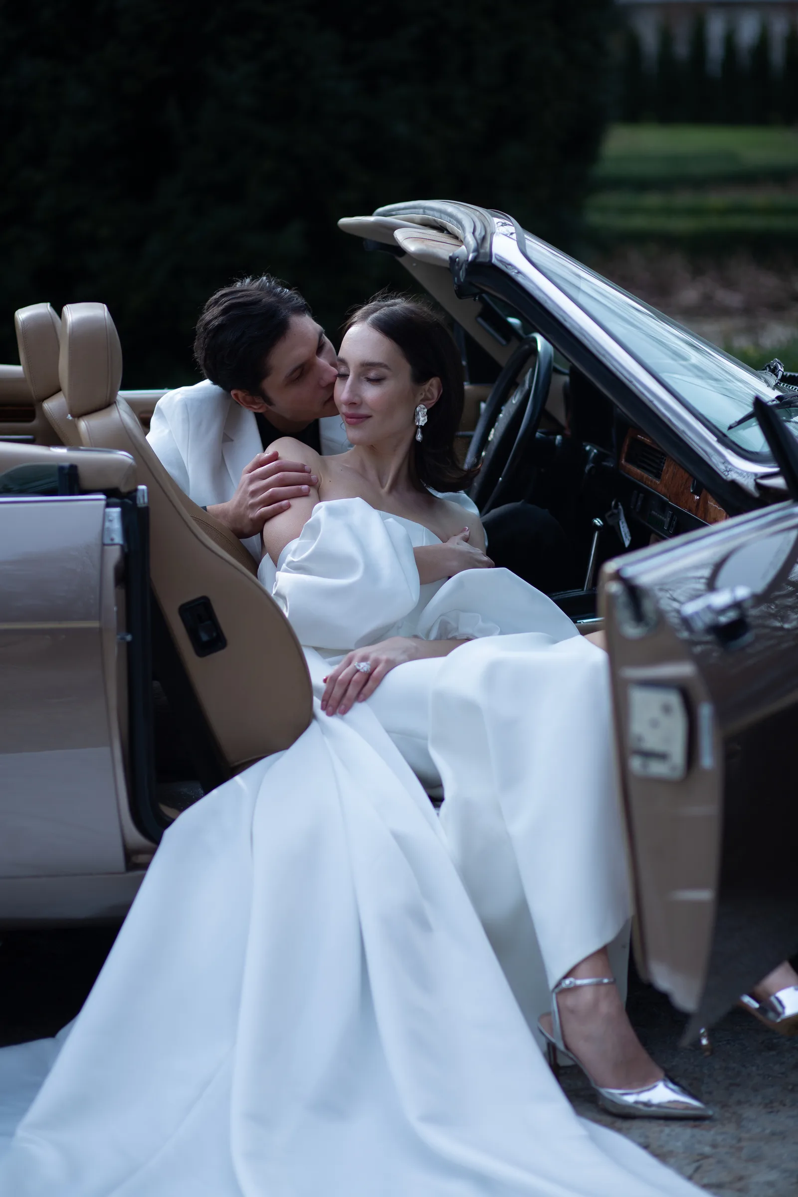 Brautpaar in Cabrio mit beigem Ledersitzbezug, Braut in weißem Seidenkleid, klassisches Hochzeitsfoto