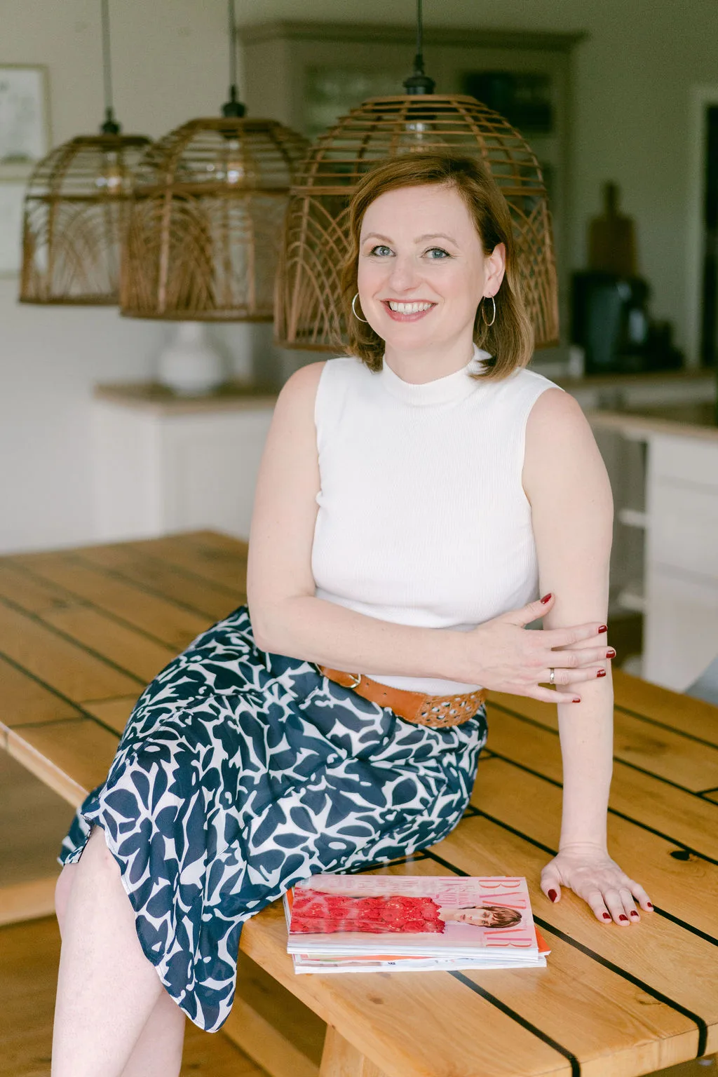 Presse Rockstars – PR-Coaching für Medienpräsenz von Marike Frick 11 Frau mit braunem Haar in weißem Top und gemustertem Rock sitzt auf Holzbank vor Rattan-Pendelleuchten