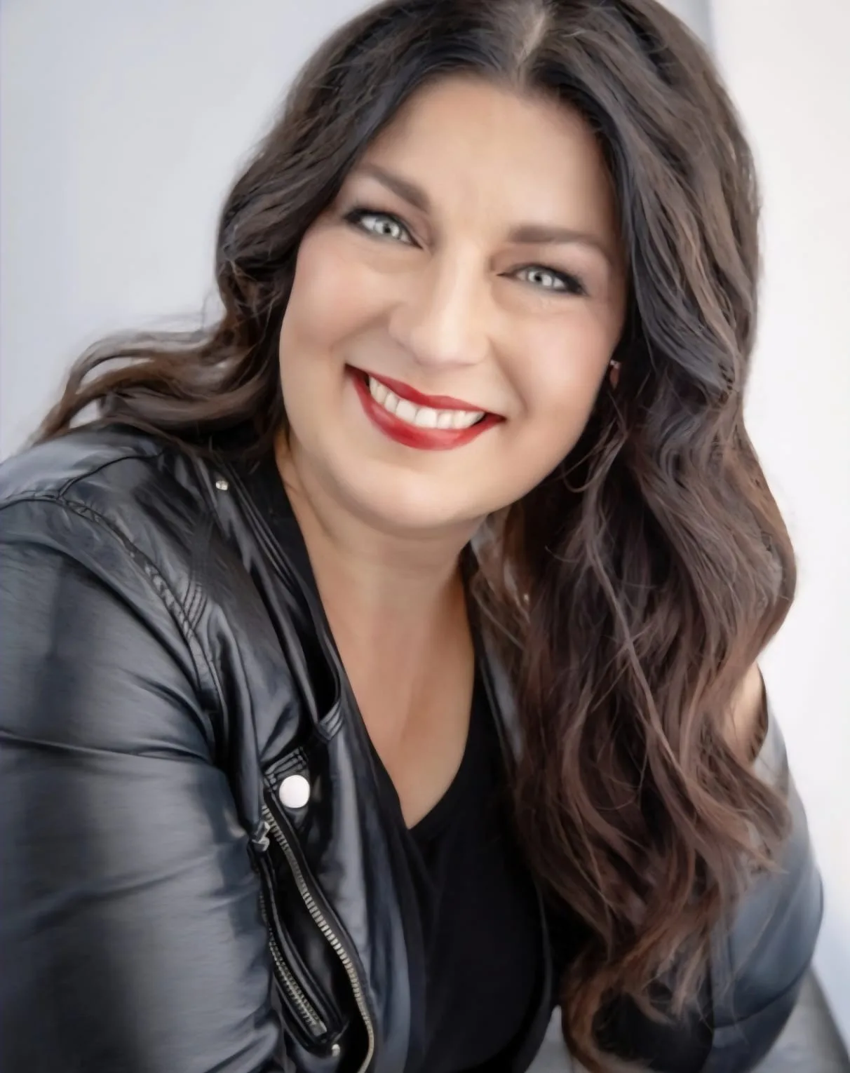 Frau mit langen braunen Haaren, rotem Lippenstift, schwarzer Lederjacke, lächelnd vor grauem Hintergrund
