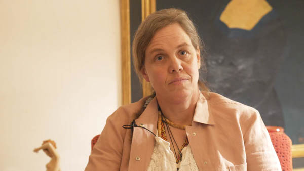 Melanie H., sitzt auf Couch in brauner Jacke vor hellem Hintergrund