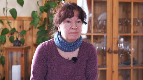 Christel W., Krankenschwester, in violettem Pullover und blauem Schal vor Bücherregal