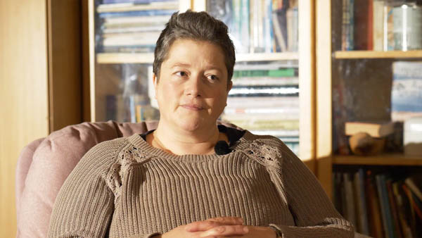 Natalie H., Pflegerin, in beigem Pullover sitzend vor Bücherregal