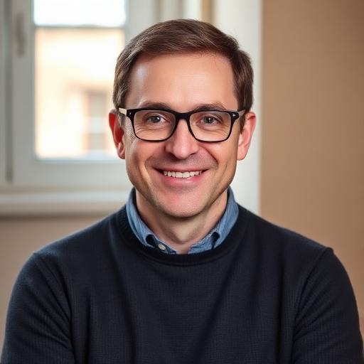 Profilbild eines lächelnden Mannes mit dunklen Haaren und Brille im schwarzen Pullover vor hellem Hintergrund