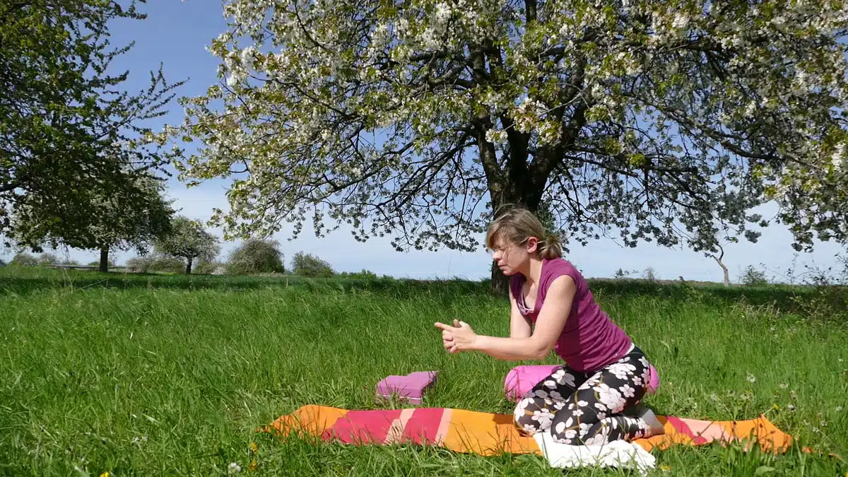 Frau sitzt in Qigong-Position auf buntem Tuch unter Kirschbaum