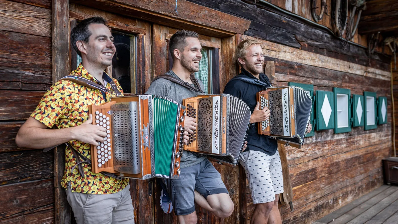 Drei Männer mit Steirischen Harmonikas vor Holzhaus-Fassade, einer in gelbem Hemd
