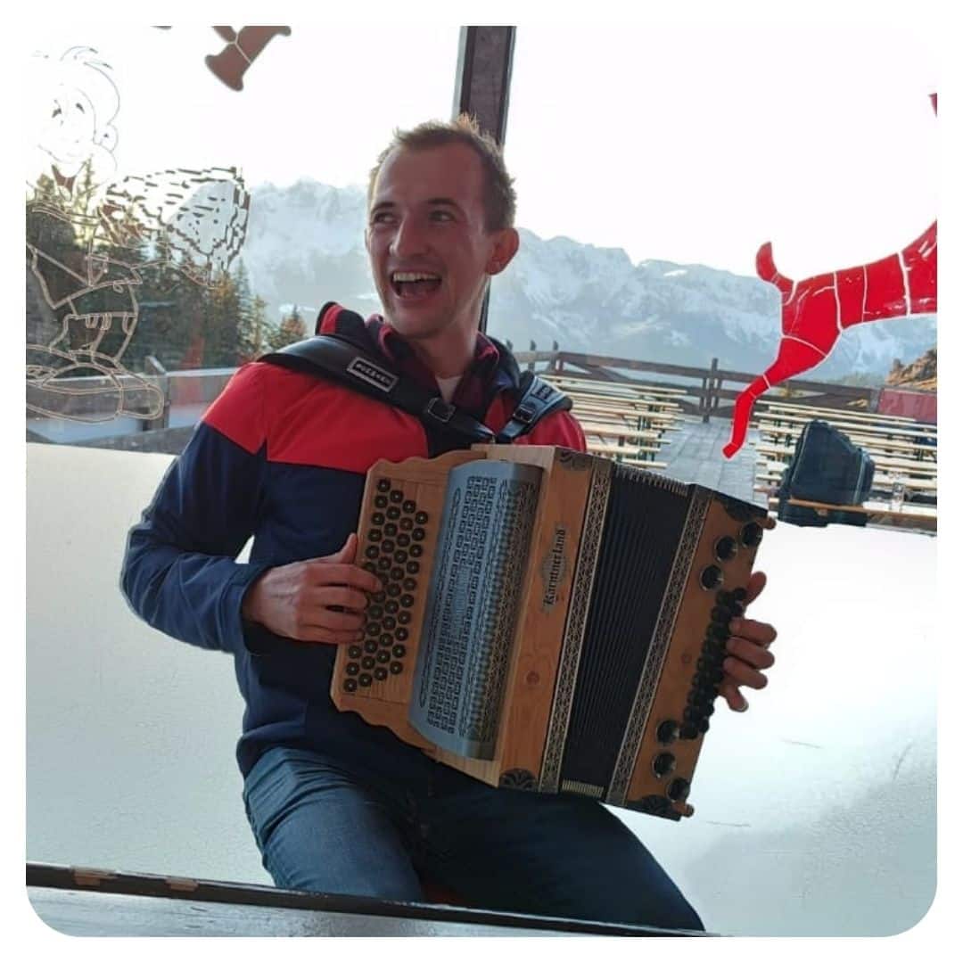 Mann mit Steirischer Harmonika auf Balkon mit Bergblick im Hintergrund