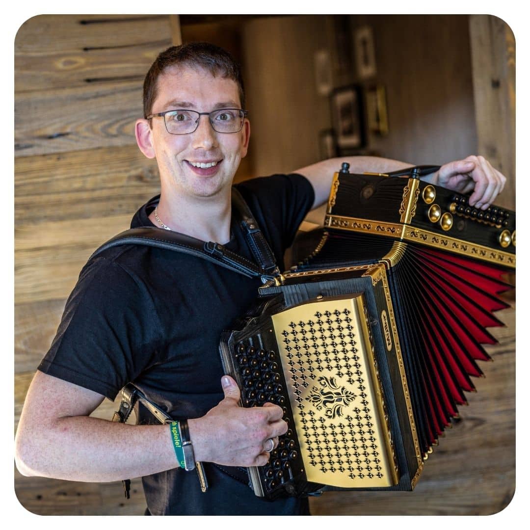 Junger Mann mit Brille hält Steirische Harmonika, schwarzes T-Shirt, lächelnd