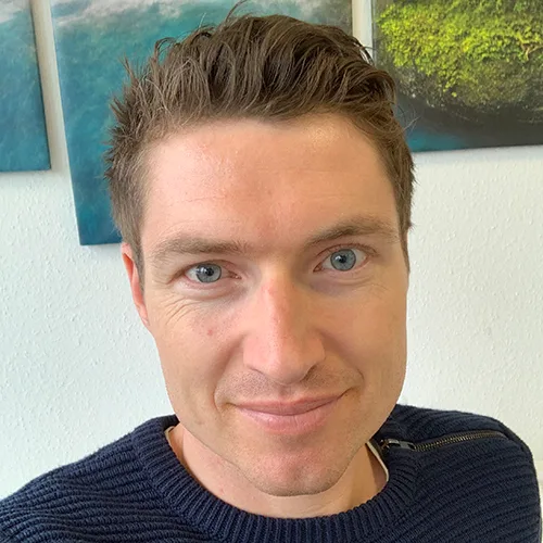 Porträt eines lächelnden Mannes mit braunen Haaren und blauem Hemd vor strukturiertem Hintergrund