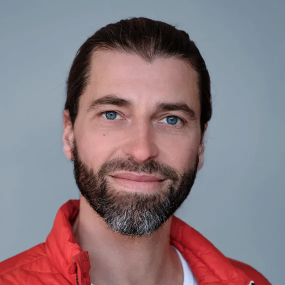 Porträt Thomas List, Mann mit Bart und dunklen Haaren in rotem Shirt vor grauem Hintergrund