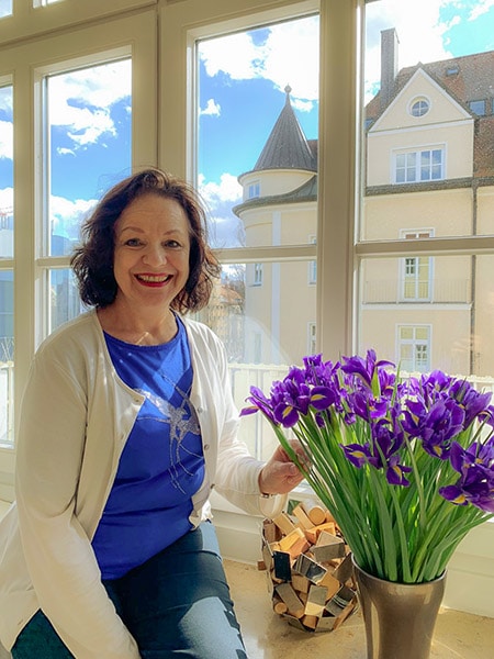 Beatrice Fischer-Stracke lächelnd am Fenster mit Blick auf Schloss, blaues Shirt, weiße Jacke, lila Irisblüten