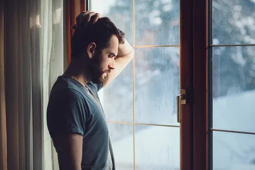 Mann sitzt nachdenklich am Fenster mit Regenwetter im Hintergrund