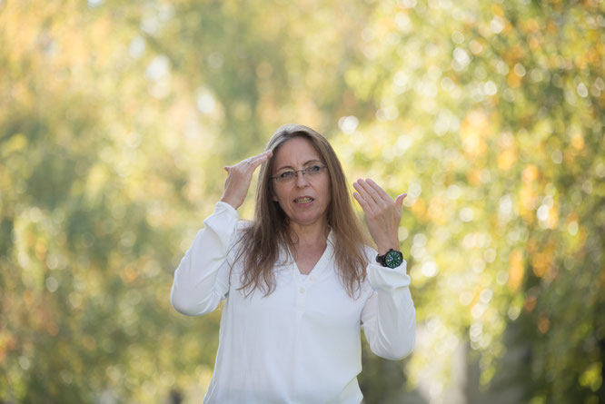 Sonja Brauner in weißem Hemd vor unscharfem grünem Laub, Hands-up-Geste