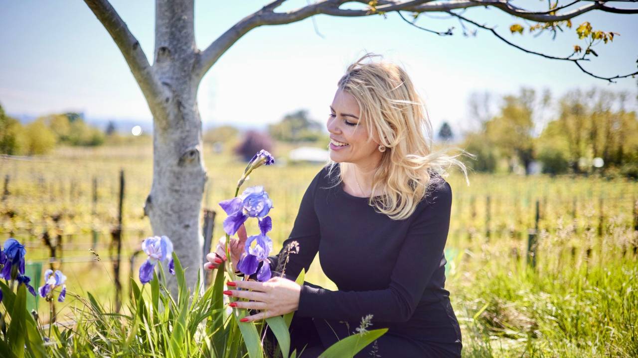 Liz Wunder mit blondem Haar lächelnd, schwarzes Oberteil, Feld mit violetten Iris-Blüten und Baum im Hintergrund