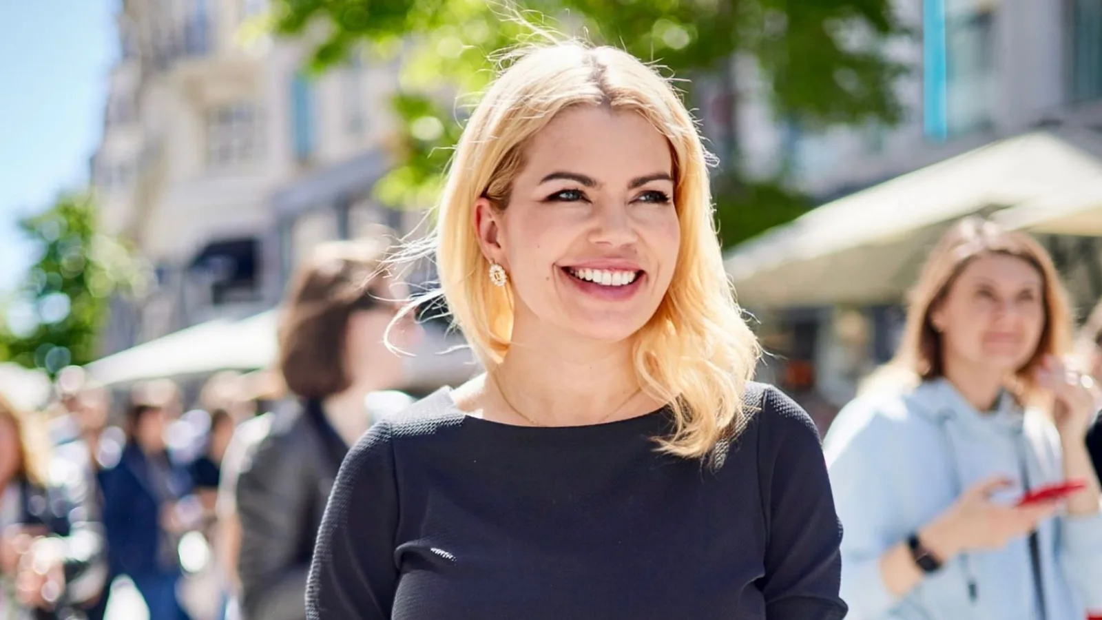 Liz Wunder mit blondem Haar lächelnd, schwarzes Oberteil, urbane Umgebung mit Terrasse und Menschen im Hintergrund