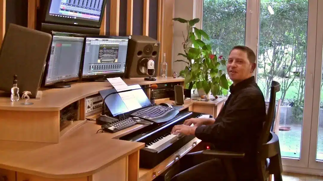 Roman Sillipp sitzt am Klavier vor Computermonitoren mit DAW-Software, schwarzes Oberteil, Studio mit Pflanzen