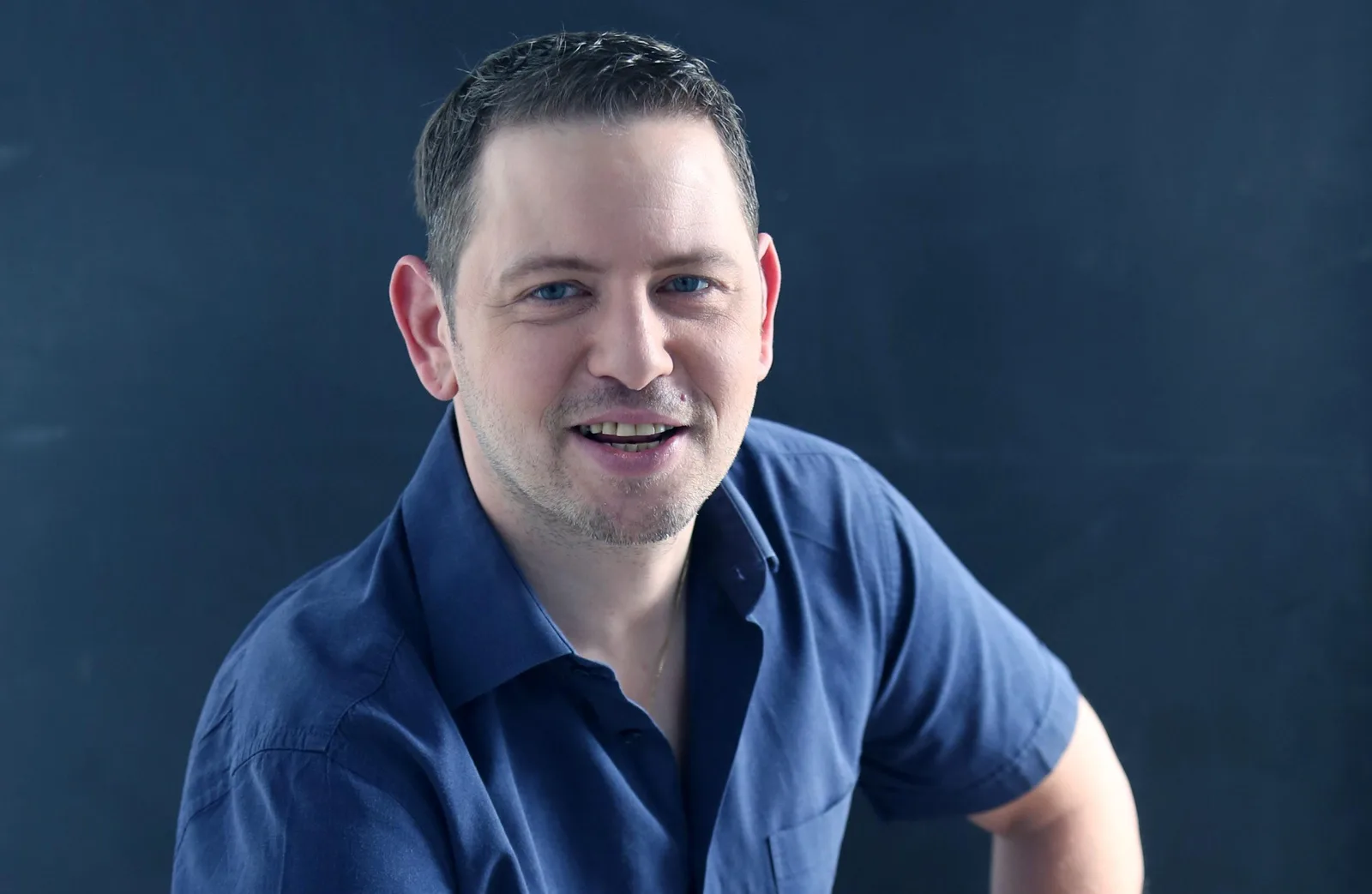 Porträt von Roman Sillipp in blauem Poloshirt vor dunklem Hintergrund, freundliches Lächeln