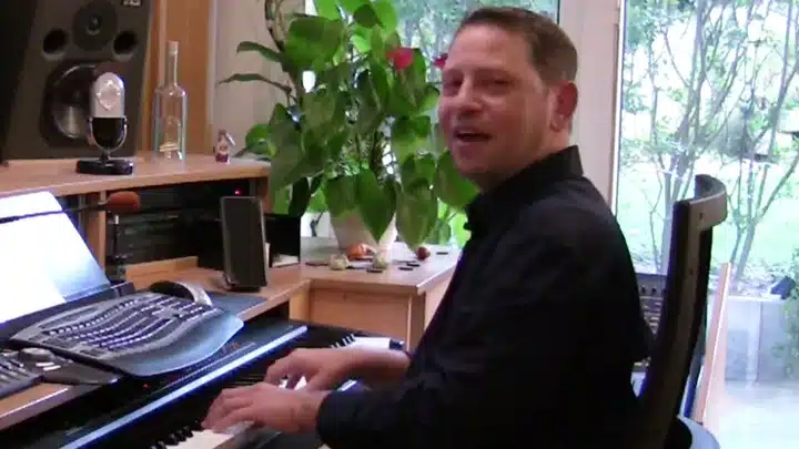 Mann sitzt am Klavier in einem Studio mit Musikequipment, Computermonitoren und Pflanze am Fenster